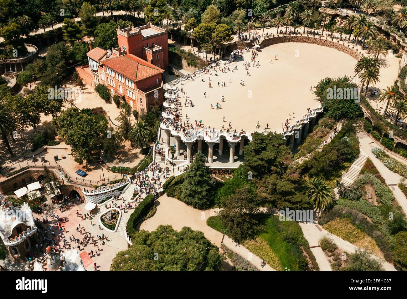 Vista aerea del Park Güell con la sua iconica terrazza piastrellata a mosaico, folle di visitatori che passeggiano sulla piazza, giardini mediterranei lussureggianti e un arco storico Foto Stock