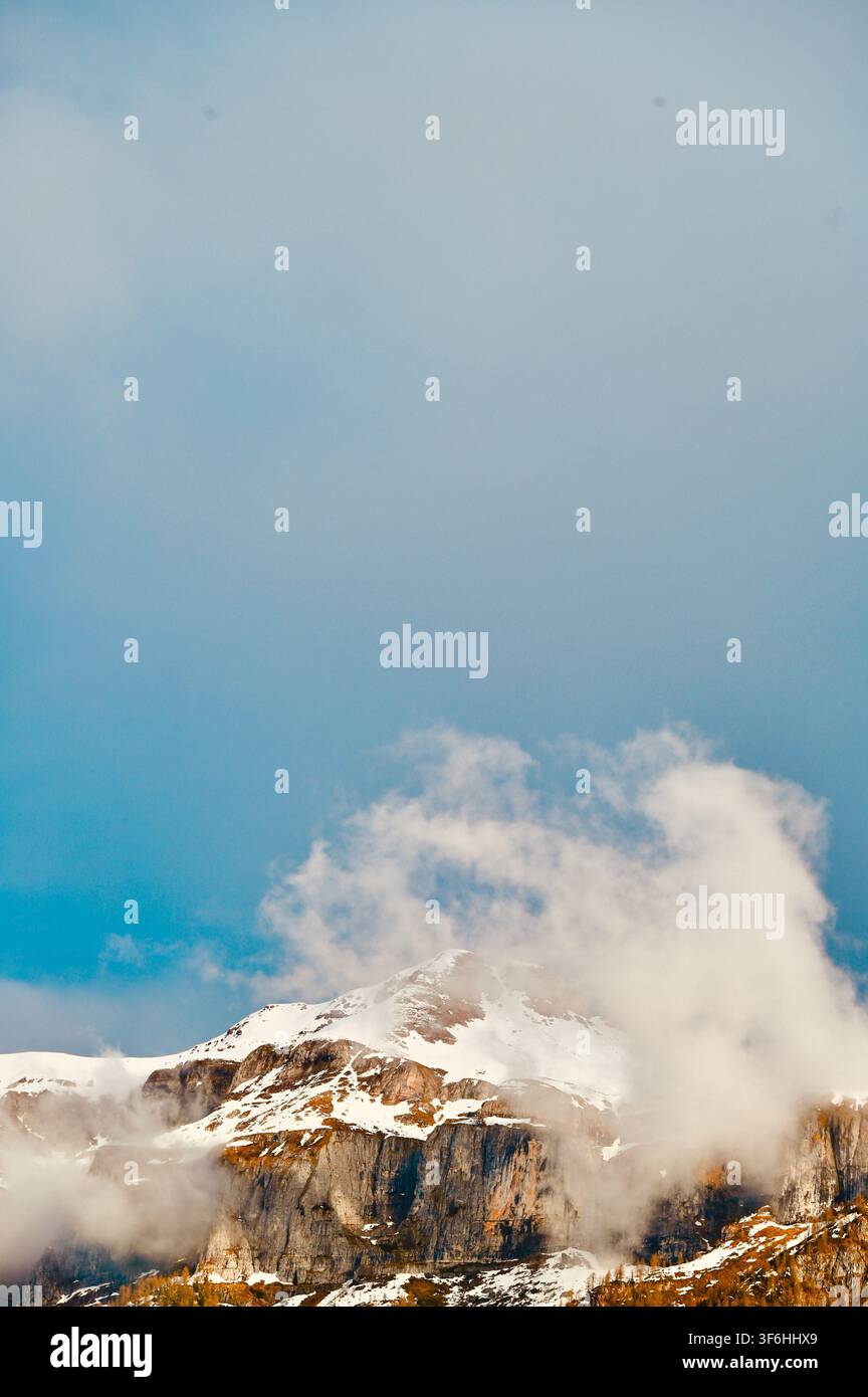 Una fotografia verticale di un maestoso picco montuoso roccioso con chiazze di neve, circondato da pendici boschive e nuvole morbide sotto un cielo azzurro. Foto Stock