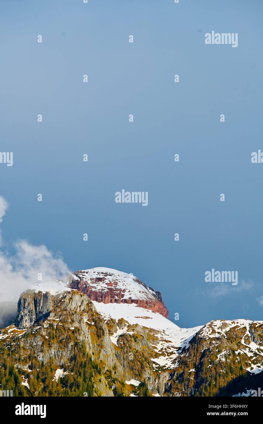 Una fotografia verticale di un maestoso picco montuoso roccioso con chiazze di neve, circondato da pendici boschive e nuvole morbide sotto un cielo azzurro. Foto Stock