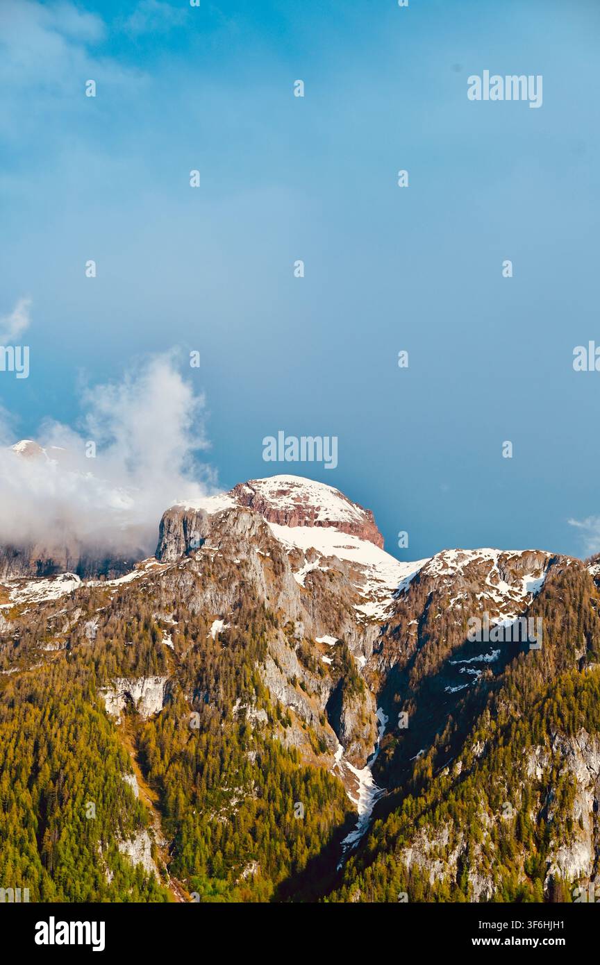 Una fotografia verticale di un maestoso picco montuoso roccioso con chiazze di neve, circondato da pendici boschive e nuvole morbide sotto un cielo azzurro. Foto Stock