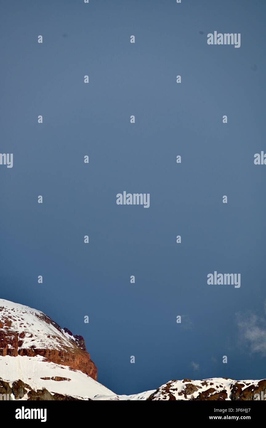 Una fotografia verticale di picchi montuosi frastagliati, ricoperti di neve, che si innalzano sopra pendici boscose sotto un cielo blu pallido. Foto Stock