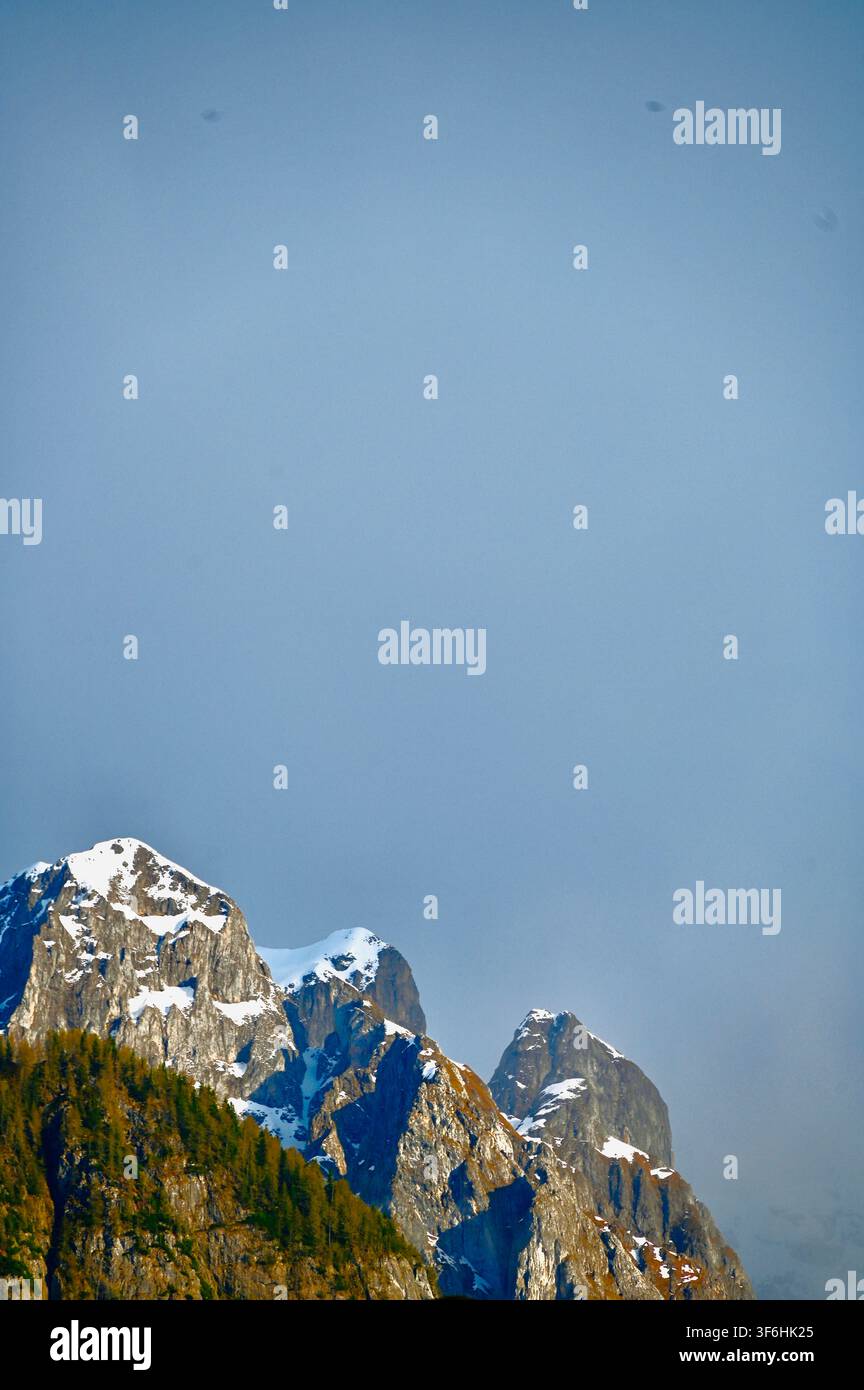 Una fotografia verticale di picchi montuosi frastagliati, ricoperti di neve, che si innalzano sopra pendici boscose sotto un cielo blu pallido. Foto Stock