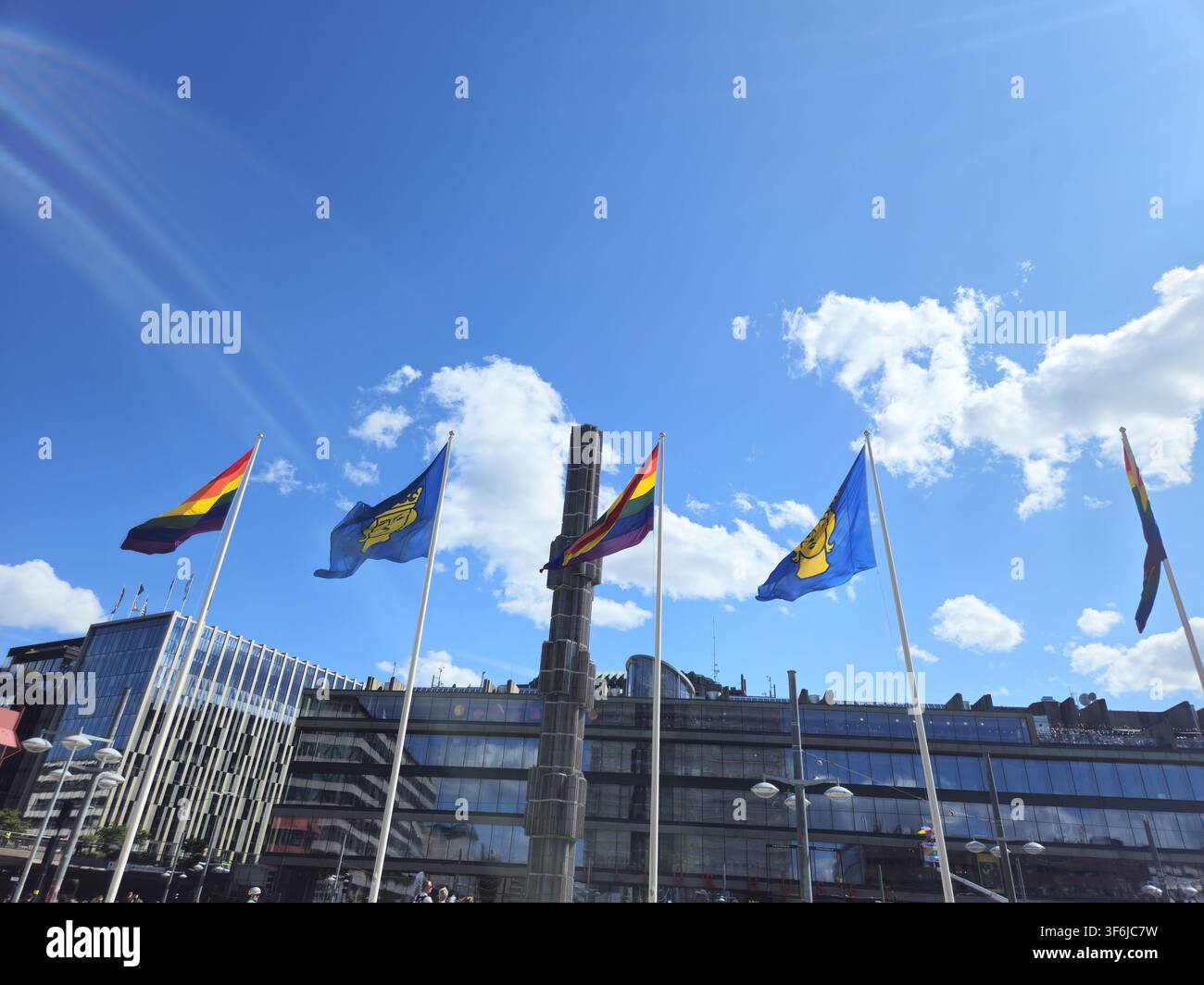 Stoccolma, Svezia, 1 agosto 2026. Bandiere arcobaleno colorate e bandiere comunali blu sventolano sopra una vivace piazza urbana con edifici moderni in vetro, br Foto Stock
