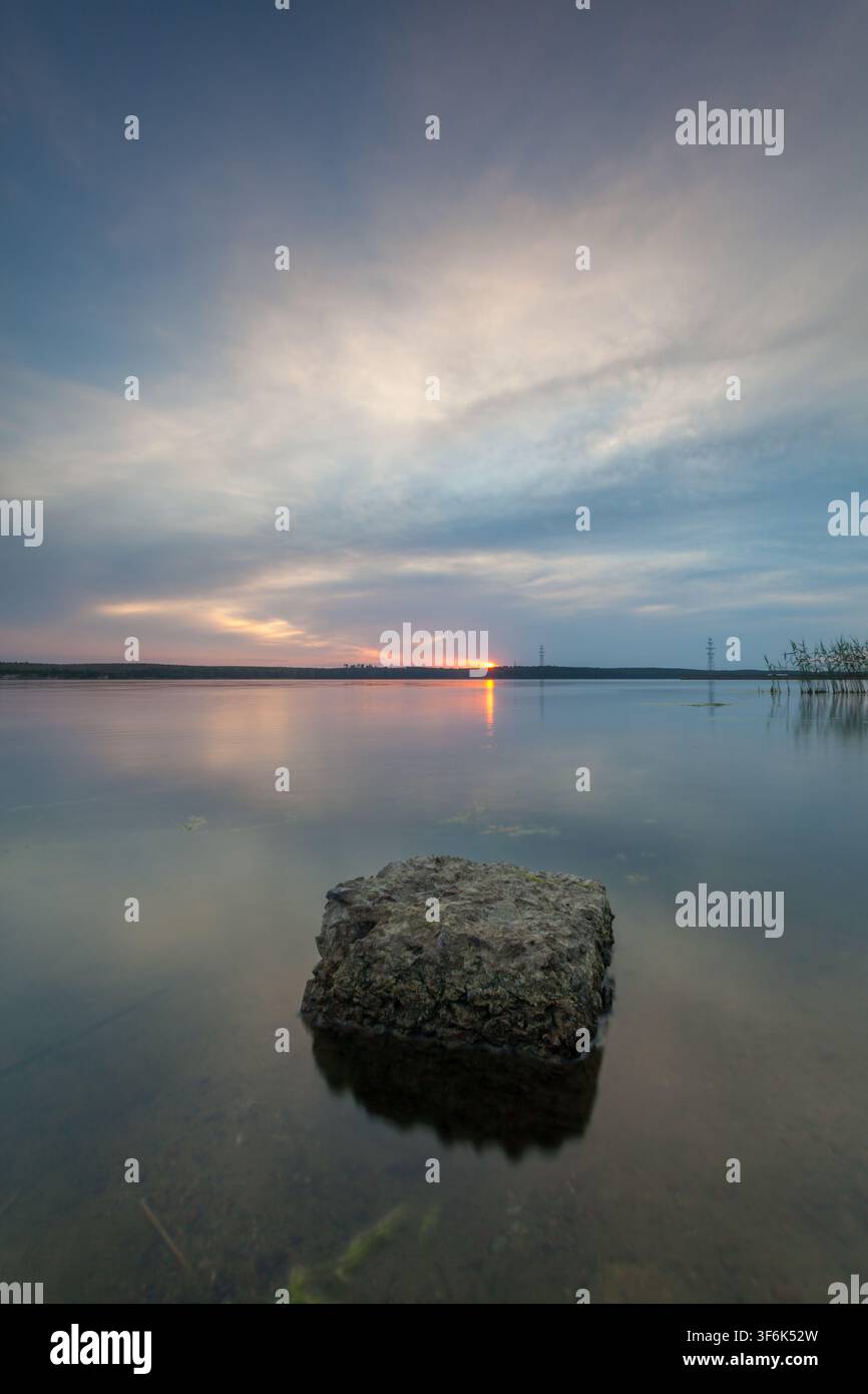 Lago calmo al tramonto con rocce nell'acqua chiara e bassa, cielo soffice e nuvoloso e canne lungo la riva. Paesaggio naturale tranquillo con riflessi e luce delicata Foto Stock