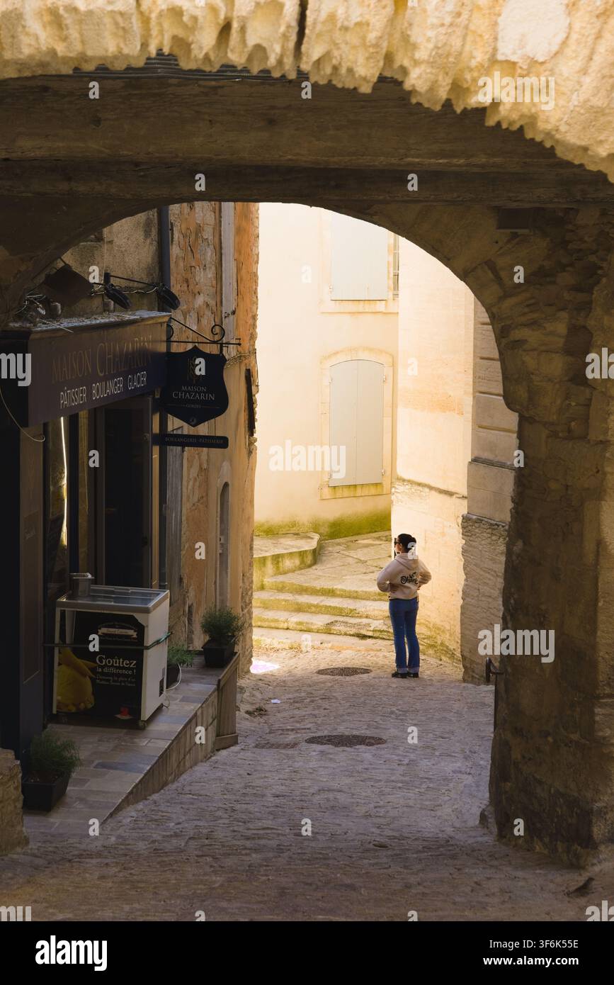 Vista attraverso un antico arco di pietra verso una panetteria e una turista in un vicolo stretto a Gordes, Francia. Foto Stock
