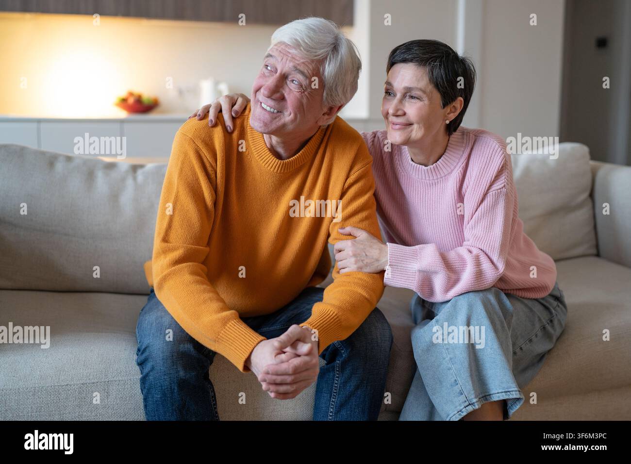 Moglie affettuosa e allegra che abbraccia il marito sorridente mentre la coppia è seduta insieme sul divano a casa. Foto Stock