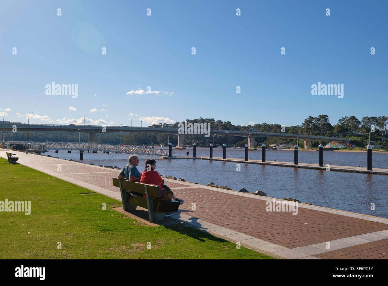 Una coppia seduta su una panchina, che si affaccia sul marciapiede in mattoni dei moli del porto, banchine. A Batemans Bay, NSW, Australia. Foto Stock