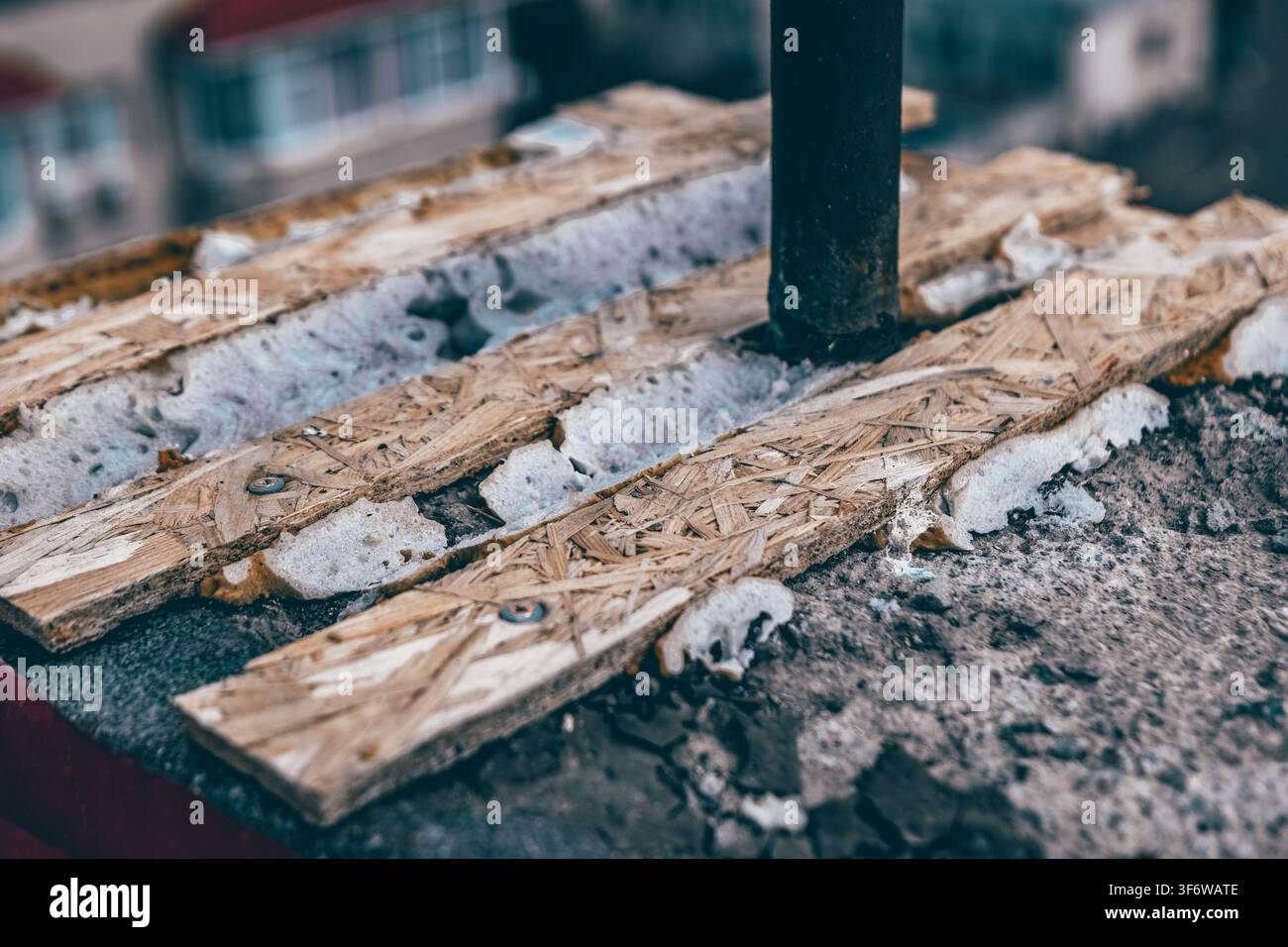Cantiere di costruzione sul tetto mostra assi di legno spaccate incastonate nel cemento sgretolato attorno a un palo metallico, con detriti e isolamento in schiuma sparsi Foto Stock