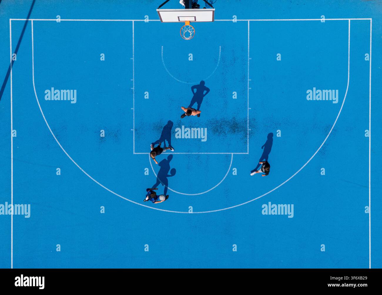 Vista aerea dall'alto di più giocatori in competizione su un campo da basket blu brillante all'aperto con lunghe ombre, concetto di gioco di basket da strada urbano. Foto Stock