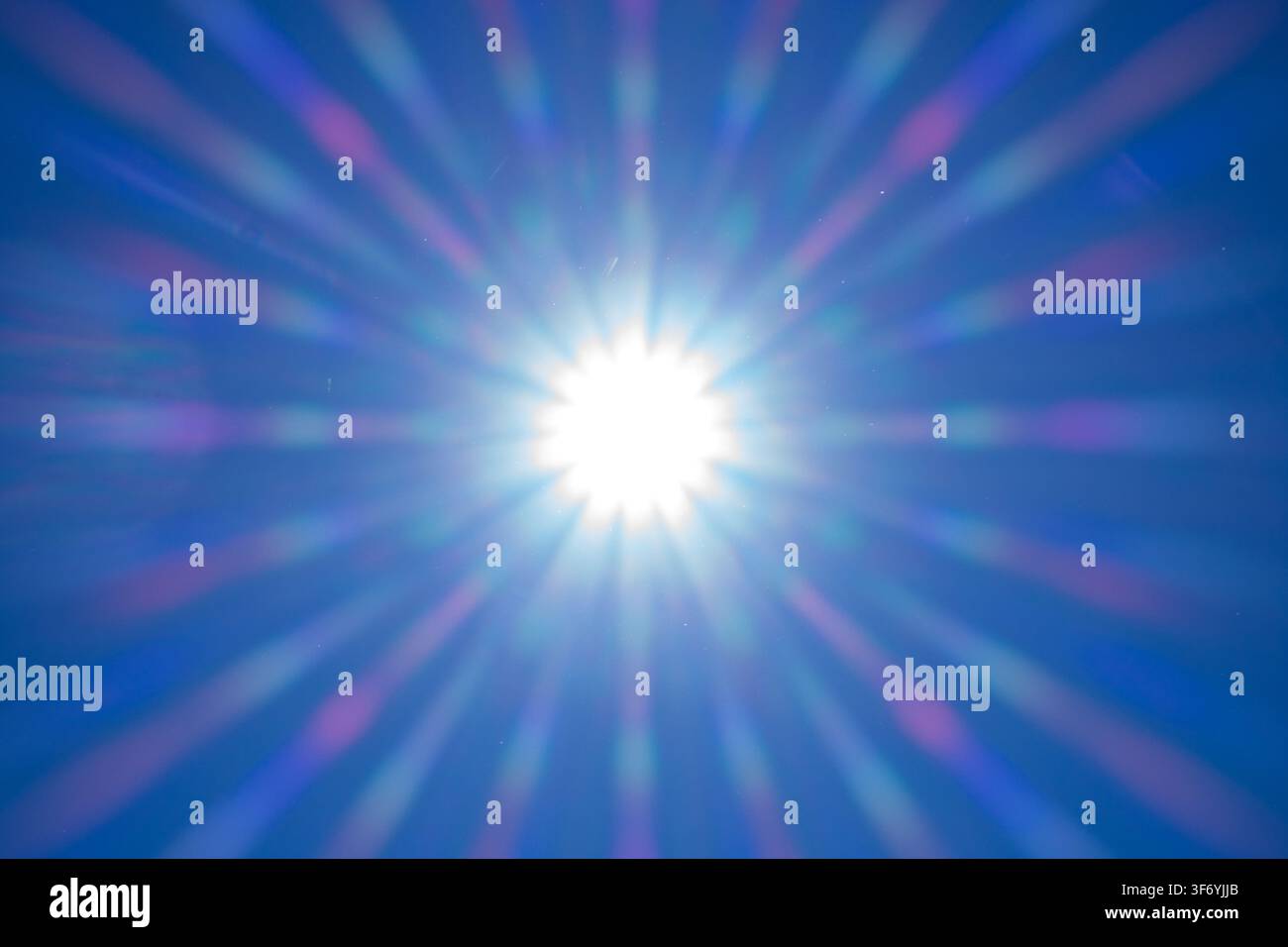 Vista diretta del sole luminoso posizionato al centro che emette raggi prismatici arcobaleno netti e simmetrici e flare ottico su un cielo blu intenso, perfe Foto Stock