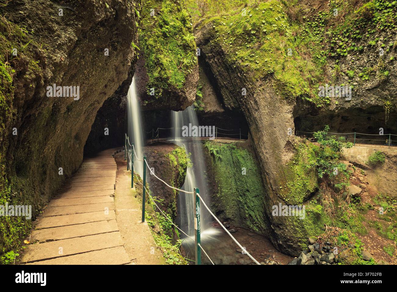 Tornadouro, Portogallo - 06.12.2025: Acqua setosa che cade da una sporgenza rocciosa ricoperta di muschio verde accanto a un marciapiede lastricato in una grotta naturale Foto Stock
