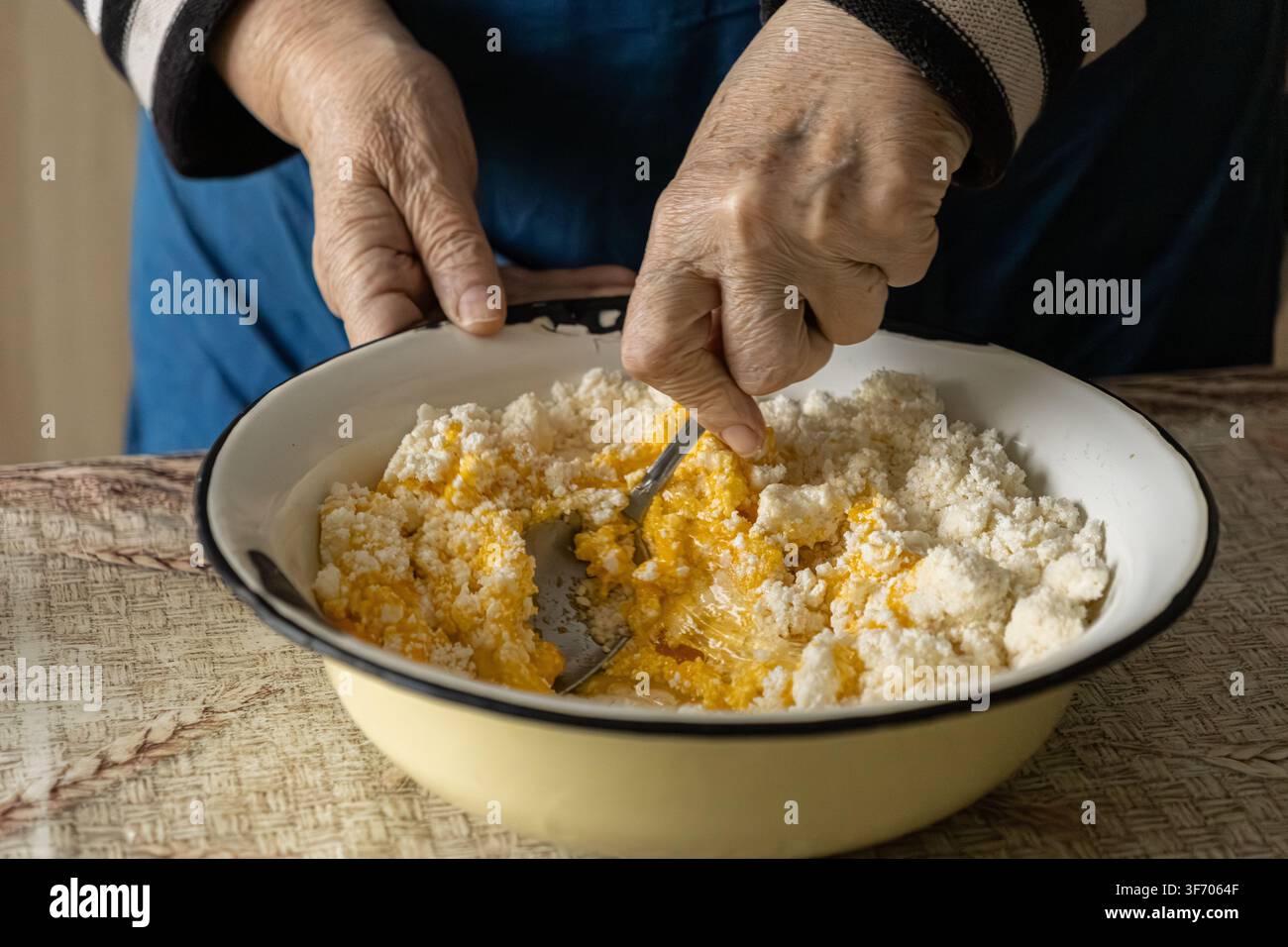 Primo piano di mani rugose che mescolano ricotta fresca con tuorli gialli in una ciotola, mostrando la preparazione dei syrniki per trasmettere una cucina autentica, eredità Foto Stock