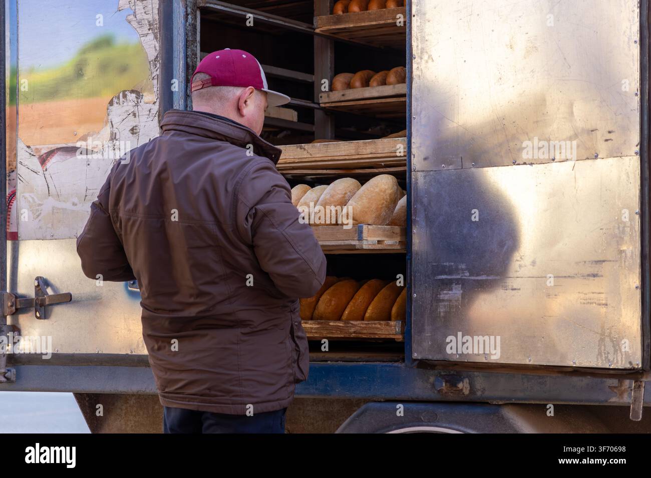 Un lavoratore rifornisce scaffali di legno all'interno di un furgone per il pane mobile, evidenziando la logistica della consegna di prodotti di panetteria freschi per le piccole imprese locali Foto Stock