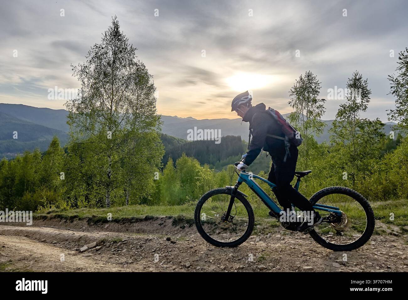 Vista laterale di un mountain biker che pedala su una bici elettrica su un sentiero sterrato attraverso colline boschive verdi al tramonto, paesaggio scenografico e concetto di avventura all'aperto Foto Stock