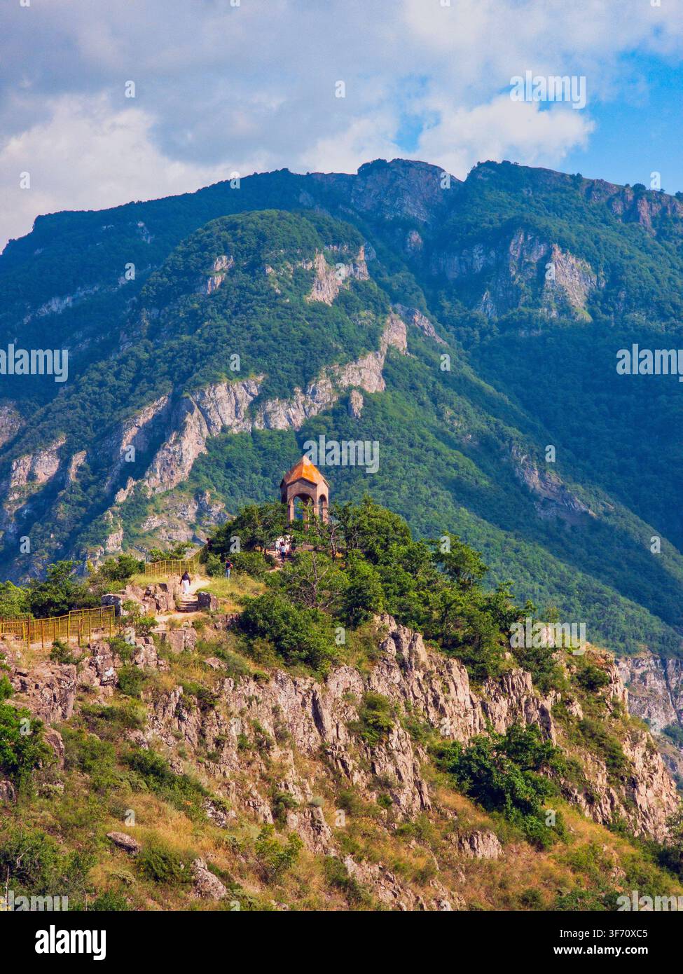 Cappella di montagna appollaiata su una scogliera rocciosa. Pendii boscosi verdi si innalzano dietro. Cielo azzurro con nuvole sparse sopra. Struttura in pietra con tetto arancione Foto Stock