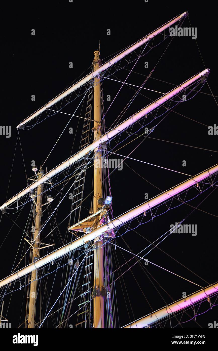 Albero di barca a vela e intricata attrezzatura con funi, aste e scale che si protendono nel cielo notturno scuro, illuminati da luci colorate Foto Stock