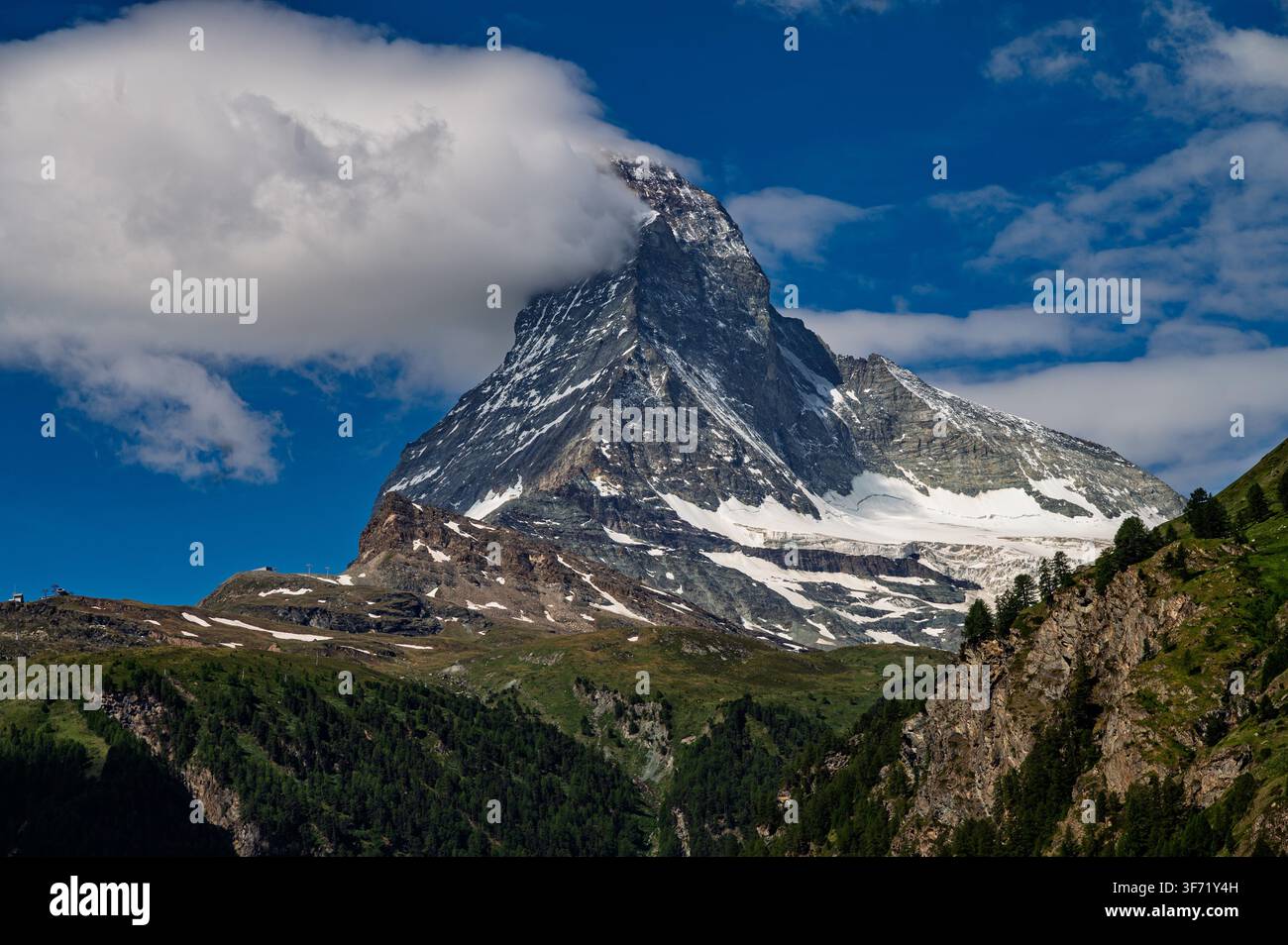 Il picco piramidale iconico del Matterhorn emerge dalle nuvole sopra i pendii alpini verdi e la foresta di pini vicino a Zermatt. Foto Stock