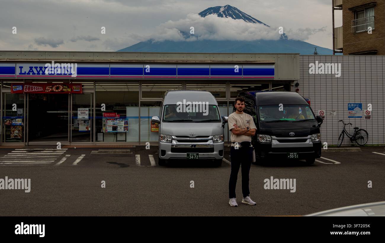 Un giovane uomo è in piedi con le braccia incrociate nel parcheggio di un minimarket con il Monte Fuji che si erge drammaticamente sullo sfondo. Foto Stock
