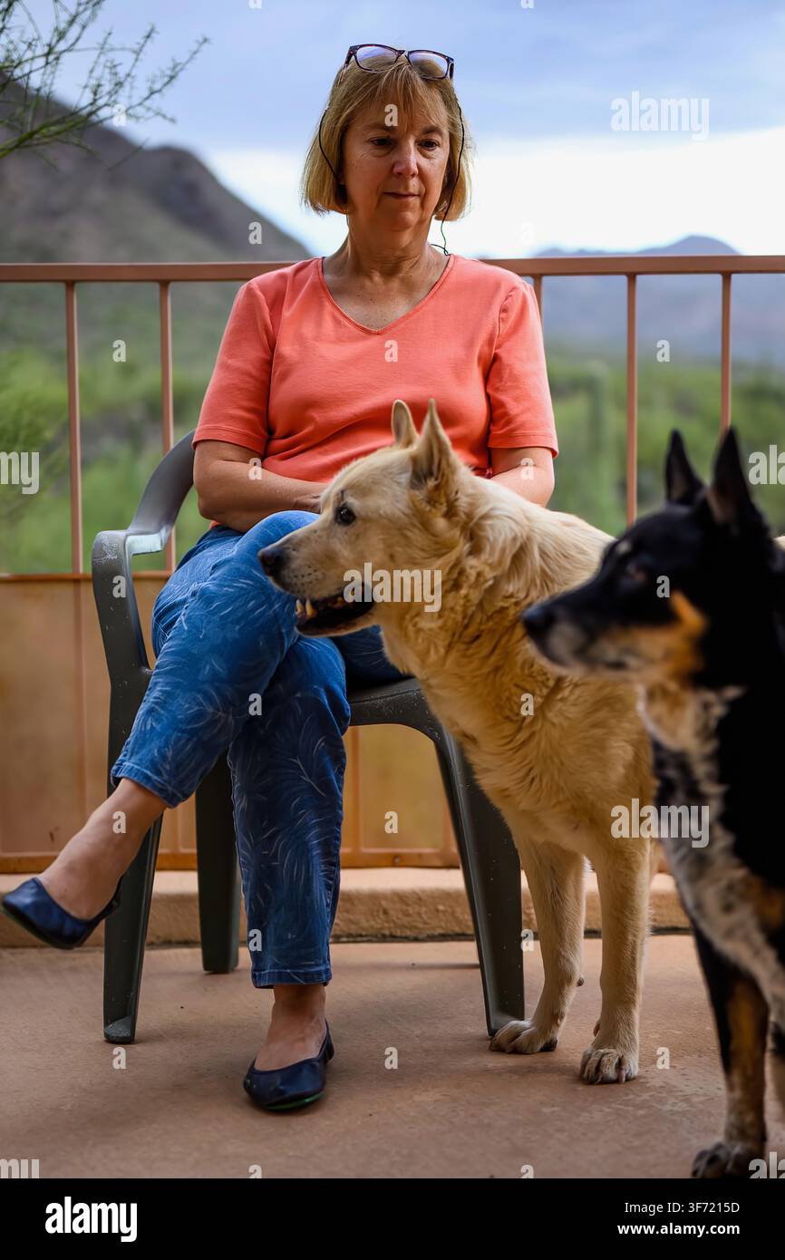 Una donna si rilassa su una sedia da patio mentre due cani, uno marrone e uno nero e marrone, stanno vicini a lei con montagne del deserto sullo sfondo. Foto Stock
