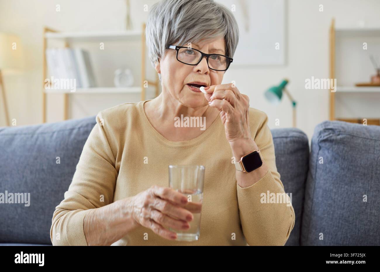 Anziana che prende pillole con acqua sul divano Foto Stock