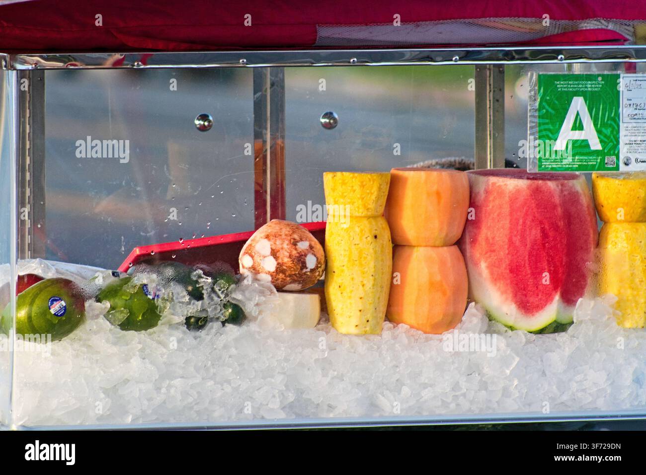 Frutta fresca si trova sopra ghiaccio tritato in un contenitore di vetro trasparente. Pezzi di anguria, melone e kiwi disposti ordinatamente. Un adesivo verde segna la qualità 'A'. Foto Stock