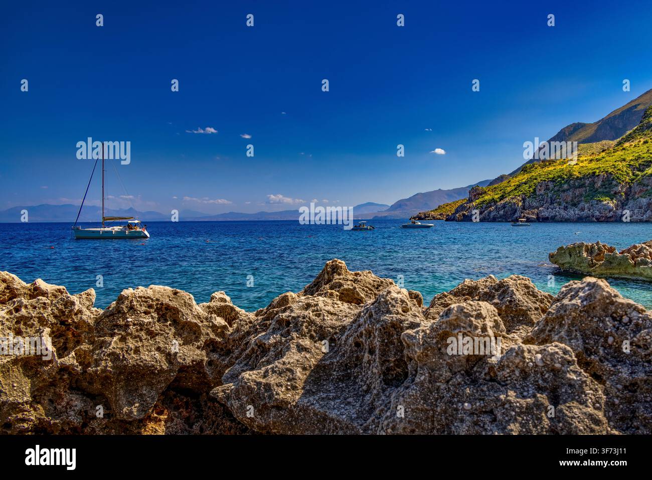Una barca a vela si posa su acque blu cristalline del Mediterraneo tra rive rocciose lungo la costa incontaminata della Riserva Naturale Orientata dello Zingaro. Foto Stock