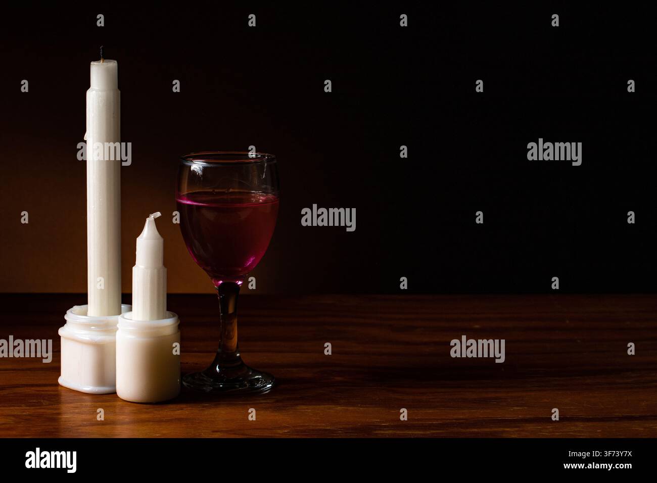 Natura morta di due candele bianche dritte non accese, con un bicchiere di vino, su una superficie a venatura del legno, con sfondo scuro ombroso dietro. Foto Stock