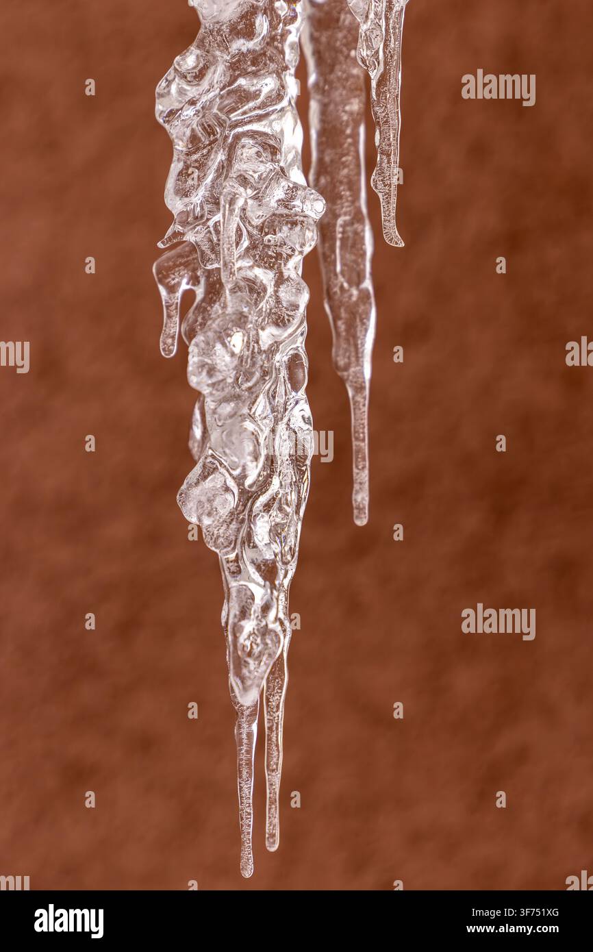Punte di ghiaccio affilate appese su uno sfondo marrone strutturato, formazioni di ghiaccio traslucide con dettagli intricati creati da motivi di acqua congelata Foto Stock
