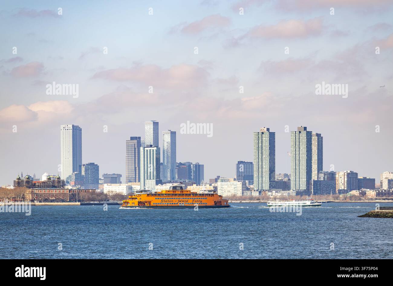 New York, USA - 20 febbraio 2026: famoso battello arancione del Staten Island Ferry che naviga sulla baia con lo skyline dei grattacieli di Jersey City sullo sfondo Foto Stock