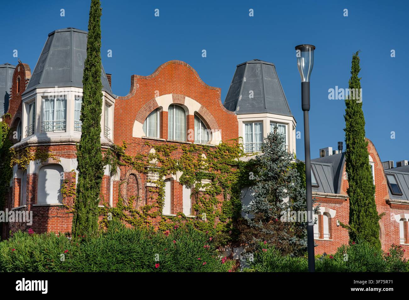 Edificio residenziale elegante in mattoni rossi con facciata ricoperta di edera e alti cipressi sotto un cielo blu Foto Stock