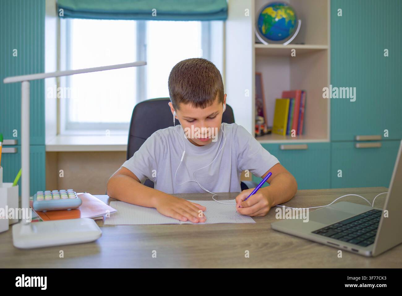 Ragazzo che scrive, in un ufficio domestico, con scrivania, laptop, blocco note, penna, da una prospettiva laterale, con accenti bianchi e blu Foto Stock