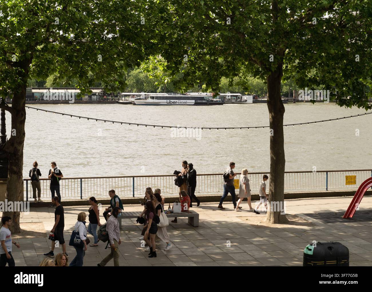 Turisti e locali passeggiano lungo il Queen's Walk sulla South Bank di Londra mentre un Uber Boat naviga lungo il Tamigi. Foto Stock