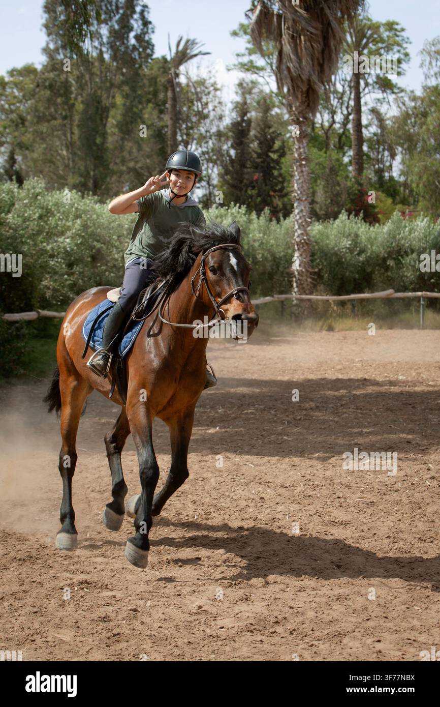 Giovane cavaliere che cavalca un cavallo marrone in un maneggio all'aperto, indossa un casco protettivo e attrezzatura equestre. Lezione di equitazione Foto Stock