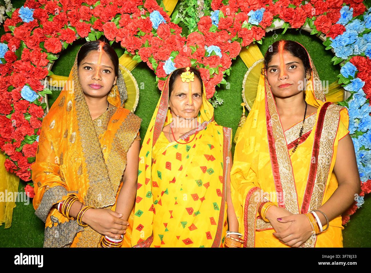 Tre donne indiane in sari gialli che posano durante una cerimonia Haldi con una decorazione a cuore floreale Foto Stock