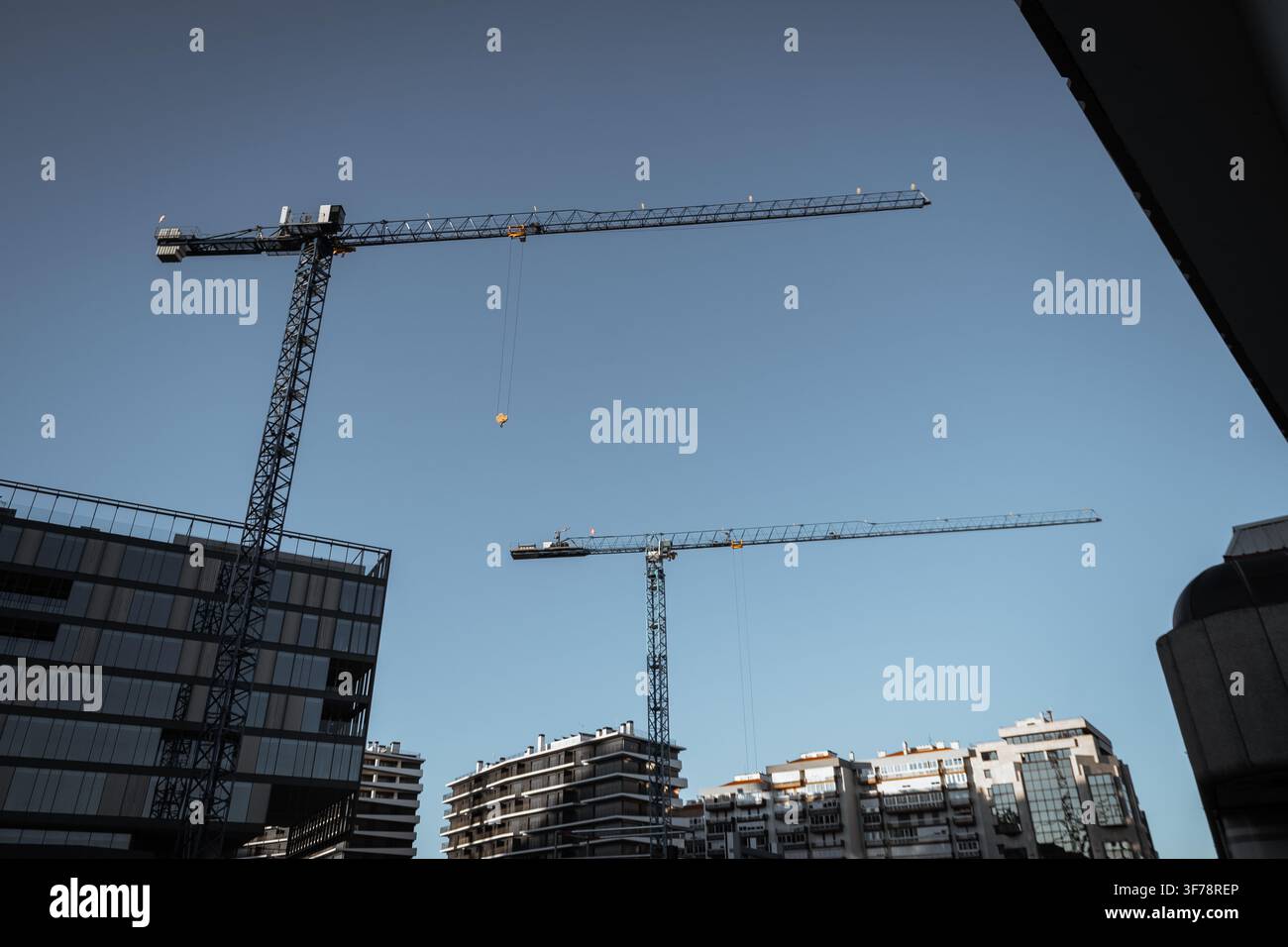 Due gru a torre si ergono sopra moderni complessi residenziali in una veduta urbana a basso angolo, illustrando la costruzione attiva, la crescita immobiliare e lo sviluppo urbano Foto Stock