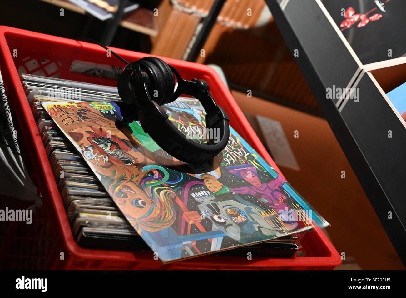 Una fotografia ad angolo alto che cattura un paio di cuffie nere appoggiate su una custodia di vinile colorata all'interno di una cassa di plastica rosso acceso piena di molti altri dischi, in un ambiente di negozio di dischi. Foto Stock
