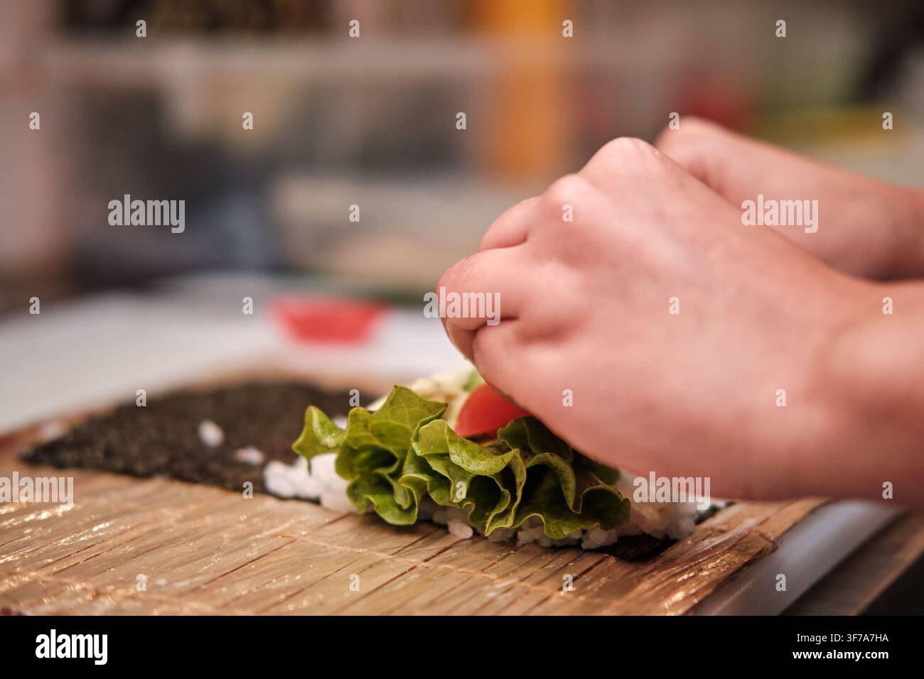 Mani che sistemano lattuga e pomodoro su riso e alghe, preparando un rotolo di sushi fresco Foto Stock