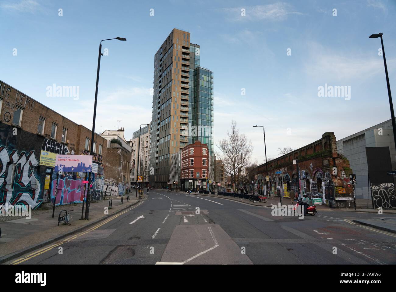 Un cavaliere fa il suo tragitto lungo Bethnal Green Road, Shoreditch, passando accanto alla torre Avant-Garde e agli archi ferroviari storici ricoperti di graffiti. Foto Stock