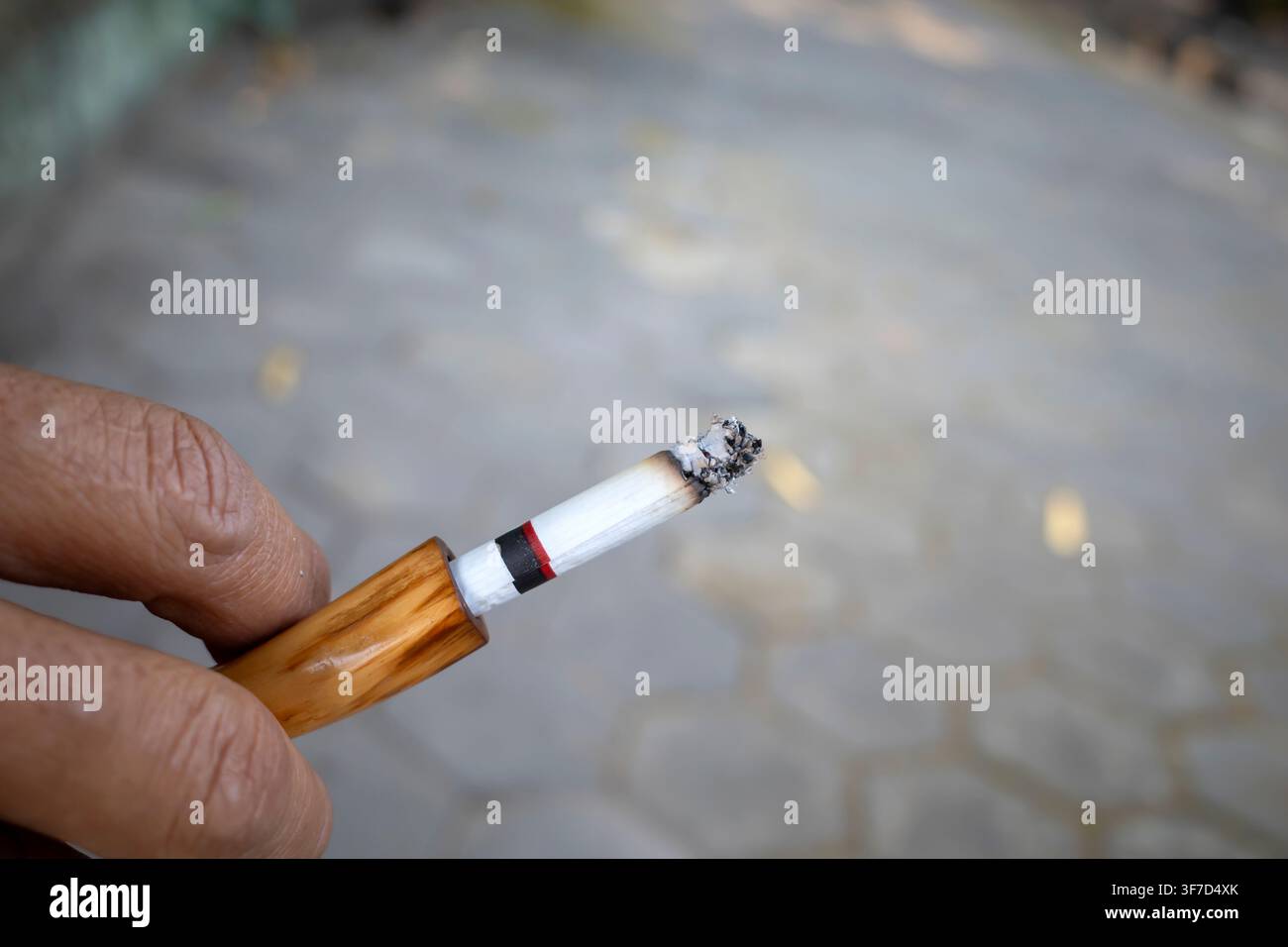 Mano che tiene una sigaretta con la punta fumante. Sfondo sfocato, mostra terreno lastricato. Focus sulla texture, cenere e fumo che si arriccia. Emozione trasmette quiete, p Foto Stock