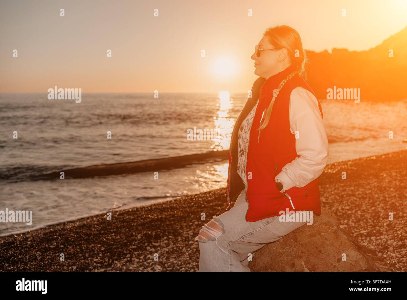 Donna seduta su una roccia in spiaggia al tramonto, indossa un giubbotto rosso e occhiali da sole mentre osserva il sole dorato sopra il mare Foto Stock
