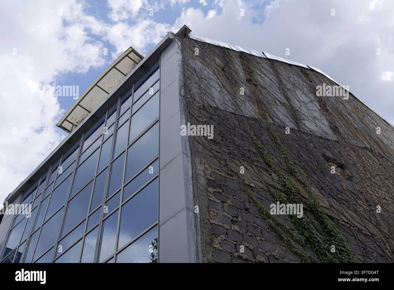 Edificio moderno con finestre di vetro riflettenti affiancato a un vecchio muro di mattoni eroso coperto da viti di edera secche, creando un forte contrasto visivo Foto Stock