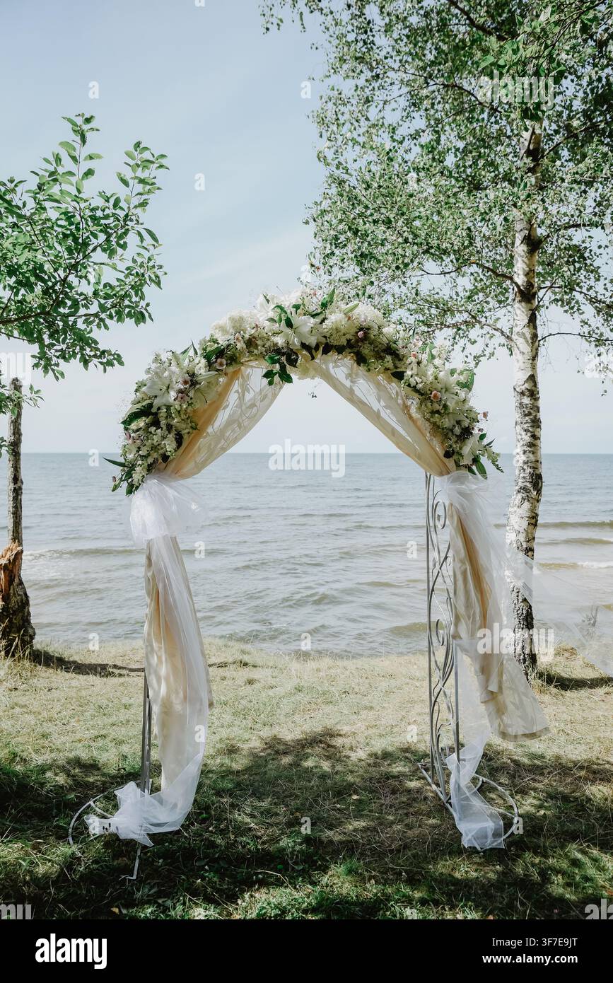 Elegante arco nuziale floreale decorato con fiori bianchi, verde e tessuto trasparente fluente, posizionato su una riva erbosa sul mare sotto le betulle, creati Foto Stock
