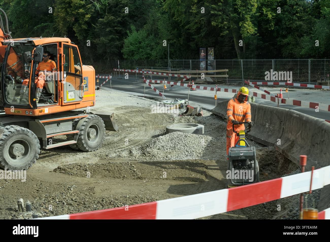 Ginevra, Svizzera – 31 luglio 2026: Due operai stradali gestiscono un escavatore e un compattatore di lastre all'interno di un cantiere stradale barricato. Foto Stock