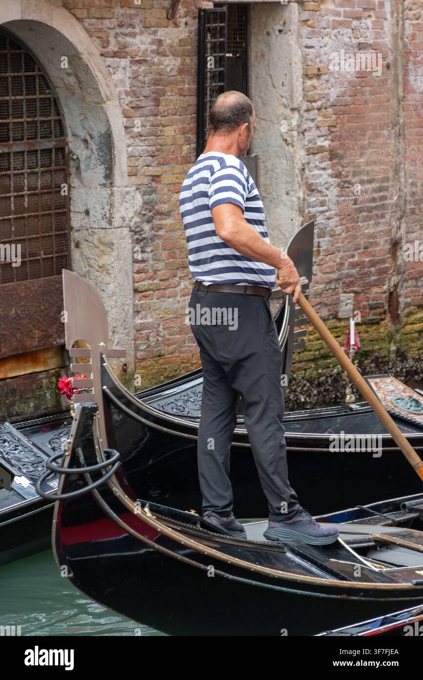 ITALIA, VENEZIA, 11 luglio 2025: Un uomo è in piedi su una gondola nera tenendo un remo vicino a un vecchio muro di canale in mattoni. Foto Stock