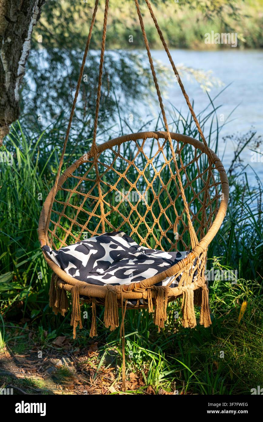 Una sedia sospesa intrecciata con un cuscino a motivi domina un fiume calmo circondato da una vegetazione lussureggiante. Ideale per il relax, la vita all'aperto, il design del giardino e il benessere Foto Stock