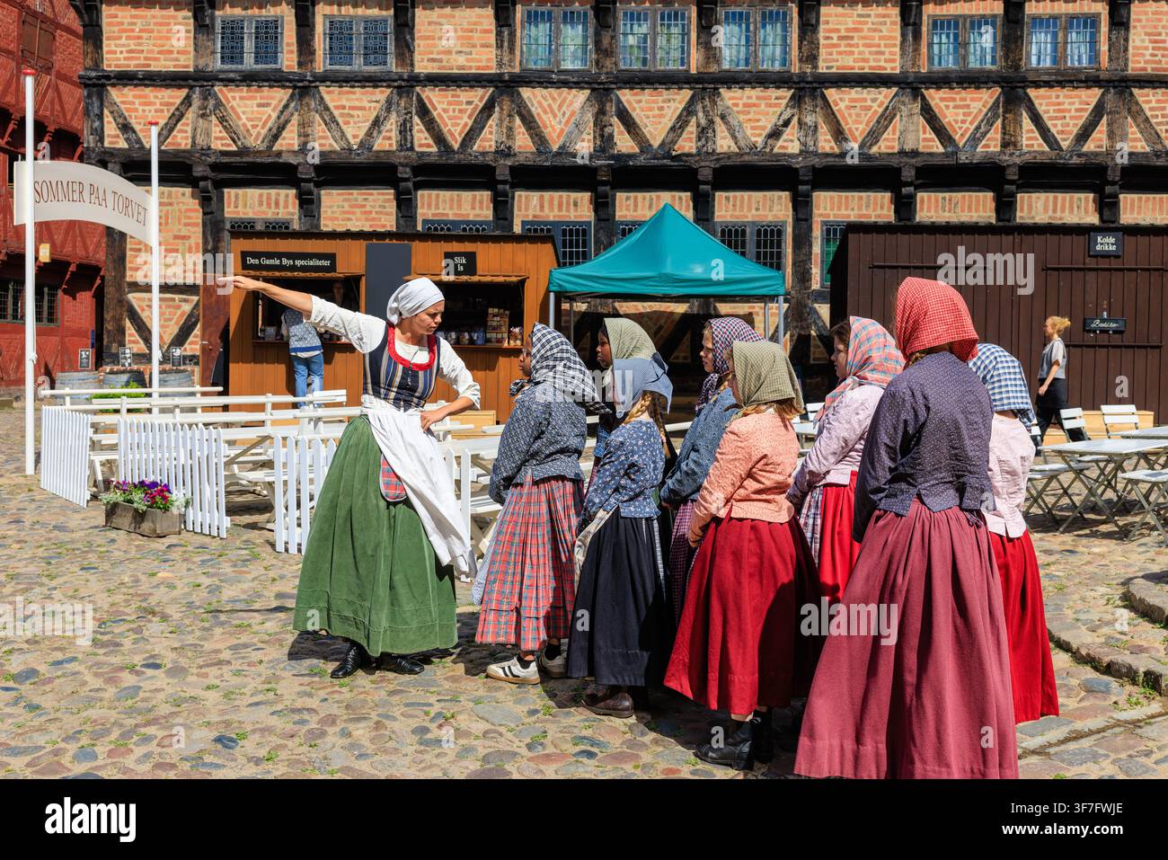 Gruppo di donne e ragazze in costumi folcloristici danesi tradizionali che ricreano una scena rurale nella storica piazza del mercato lastricata del museo Den Gamle By Foto Stock