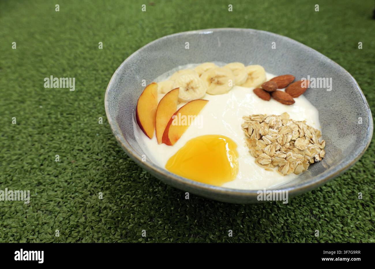 Ciotola per la colazione sana con yogurt cremoso guarnito con banane a fette, spicchi di pesche fresche, avena arrotolata, miele dolce e mandorle intere, creando un Foto Stock