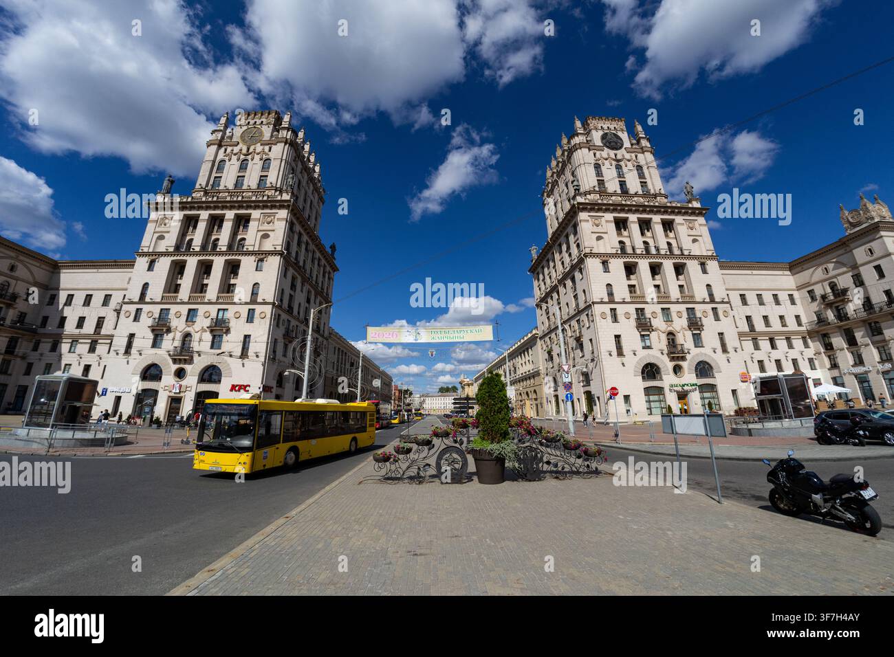 MINSK, BIELORUSSIA - 9 AGOSTO 2026: I portali iconici di Minsk (Torri Gemelle) di fronte alla stazione ferroviaria con autobus giallo sotto un cielo blu Foto Stock