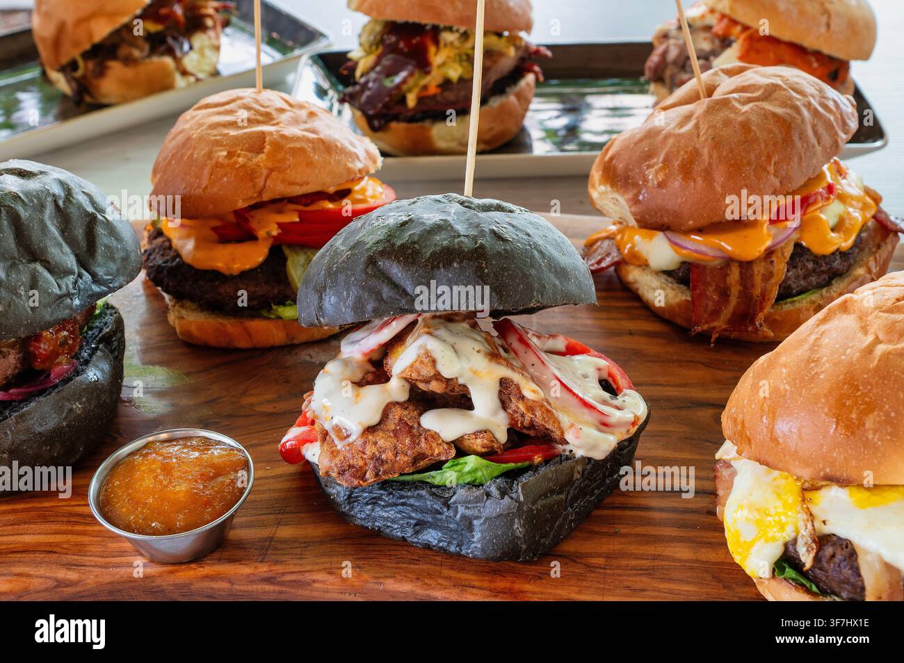 Assortimento di hamburger gourmet serviti su una tavola di legno, con panini neri e tradizionali, polpette di manzo e pollo, formaggio fuso, verdure, Bac Foto Stock