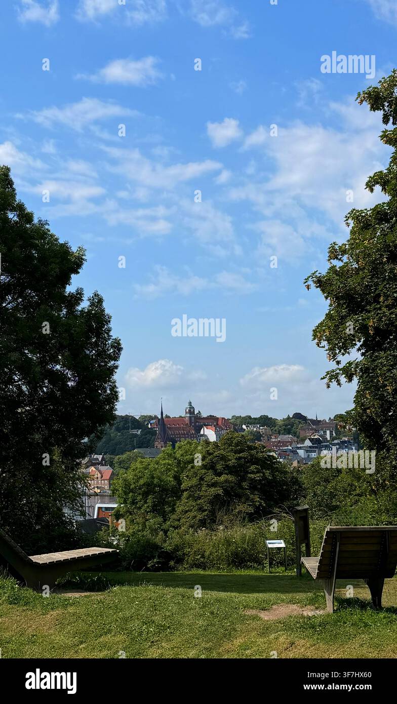 Vista panoramica di un parco verde a Flensburgo, panchina in legno che si affaccia su una skyline urbana sotto un cielo blu brillante Foto Stock